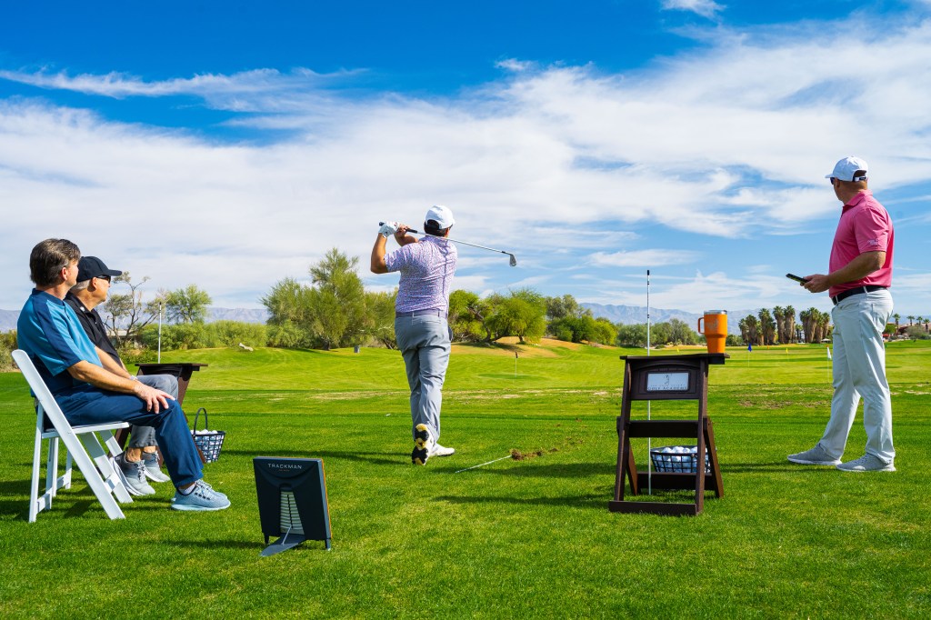 Golf student hitting an iron shot during a Palm Desert Golf Academy school session at Desert Willow Golf Resort, with instructors and participants observing on the practice range.