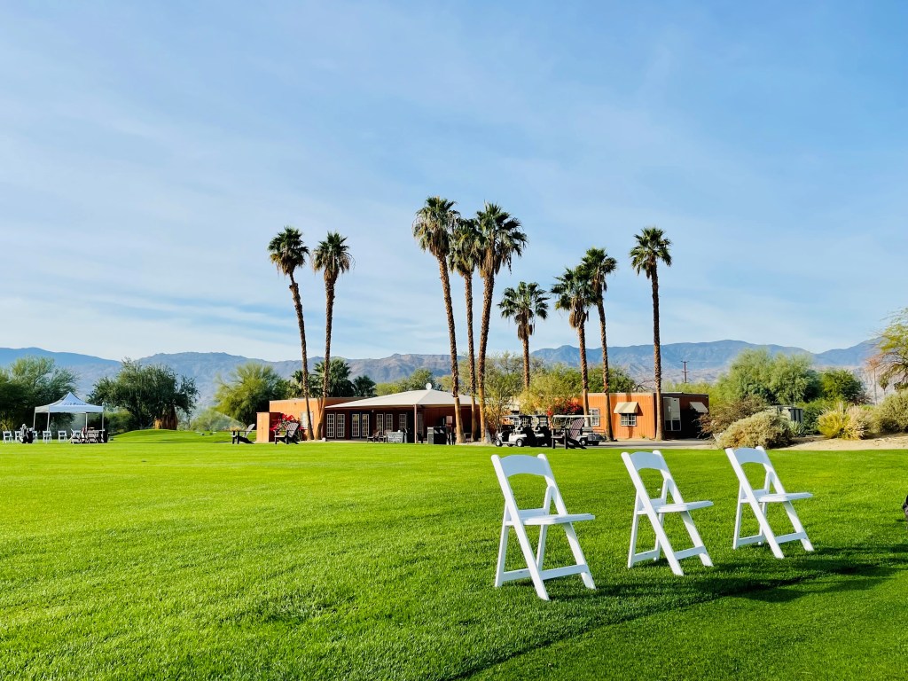 Palm Desert Golf Academy practice area set up with chairs and equipment, framed by palm trees and mountains.