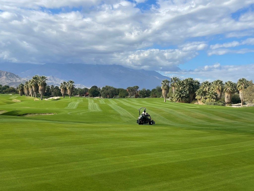 Golf course superintendent mowing lush green fairways at Desert Willow Golf Resort in Palm Desert with mountain views under a bright blue sky on Thank a Superintendent Day.