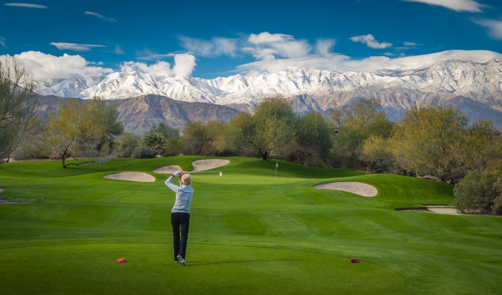 Golfer taking an on-course playing lesson at the Palm Desert Golf Academy at Desert Willow Golf Resort, with snow-capped mountains and desert landscape in the background.