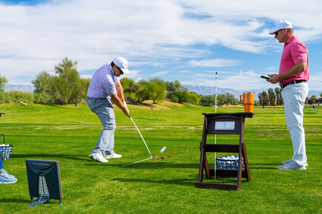 PGA Professional golf instructor coaching a student on the practice range at Palm Desert Golf Academy at Desert Willow Golf Resort, with TrackMan technology and mountain views in the background.