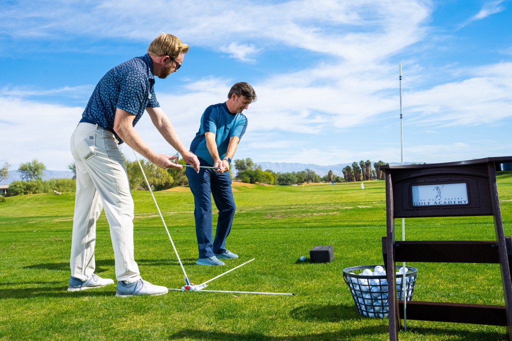PGA professional instructor providing one-on-one private golf instruction to a student at the Palm Desert Golf Academy practice range, with training equipment and golf balls nearby.