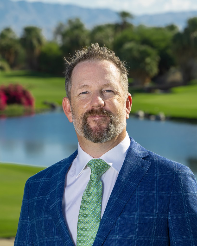Paul Bucy, PGA Professional and Director of Instruction at Palm Desert Golf Academy, headshot portrait in a suit and tie with golf course background.