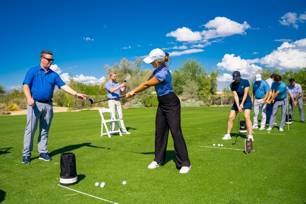 Golf instructor guiding a beginner during the Get Golf Ready program at Palm Desert Golf Academy, with other students practicing on the driving range under clear desert skies.