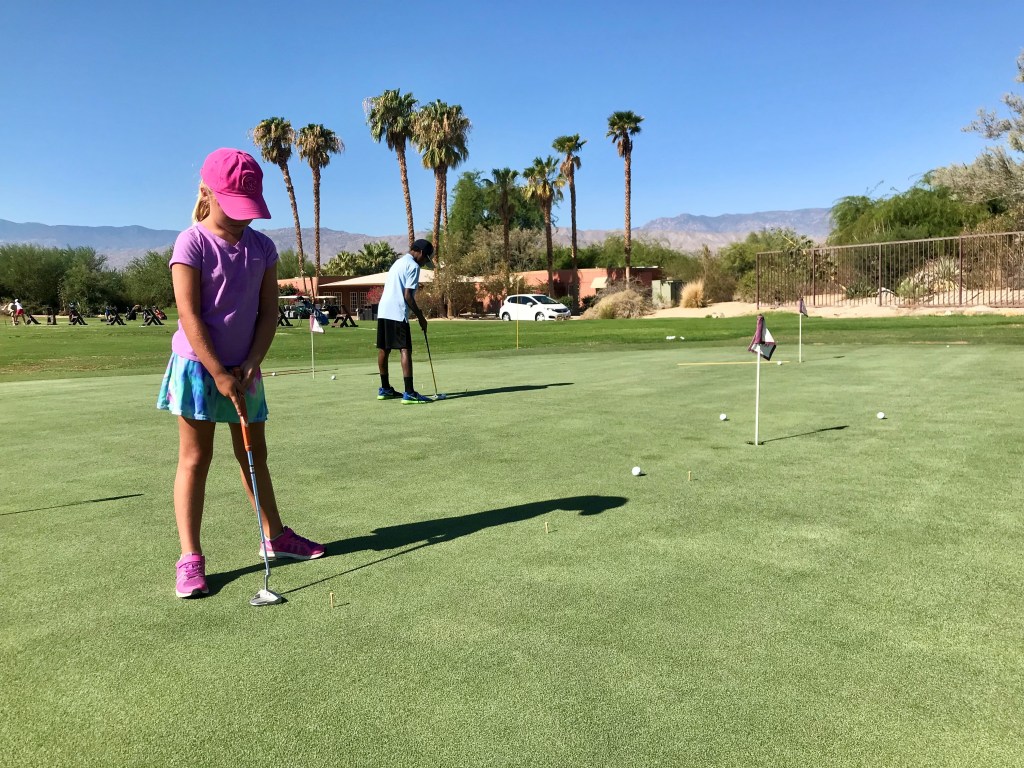 Young girl in a pink hat and purple shirt practicing putting on a sunny day at Desert Willow Golf Resort's Palm Desert Golf Academy, with palm trees and mountains in the background during the Fairway Kids Skills Series program.