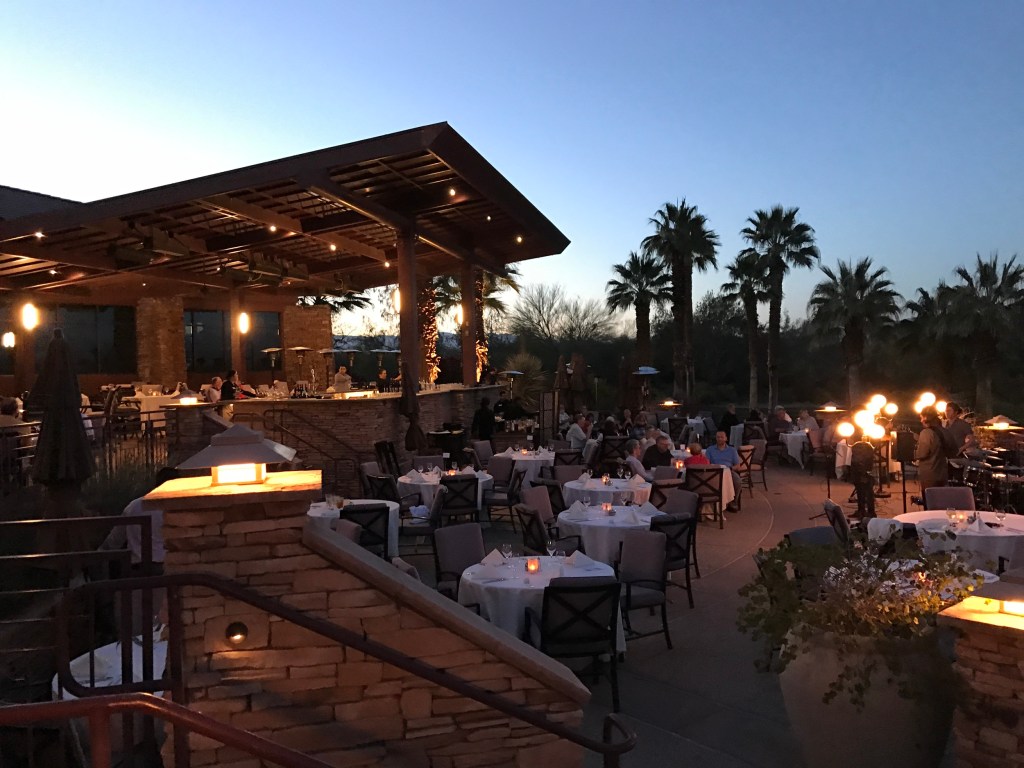Evening alfresco dining setup on the Lakeview Terrace at Desert Willow Golf Resort, with glowing lights, palm trees, and guests enjoying Music & Dining Under the Stars.