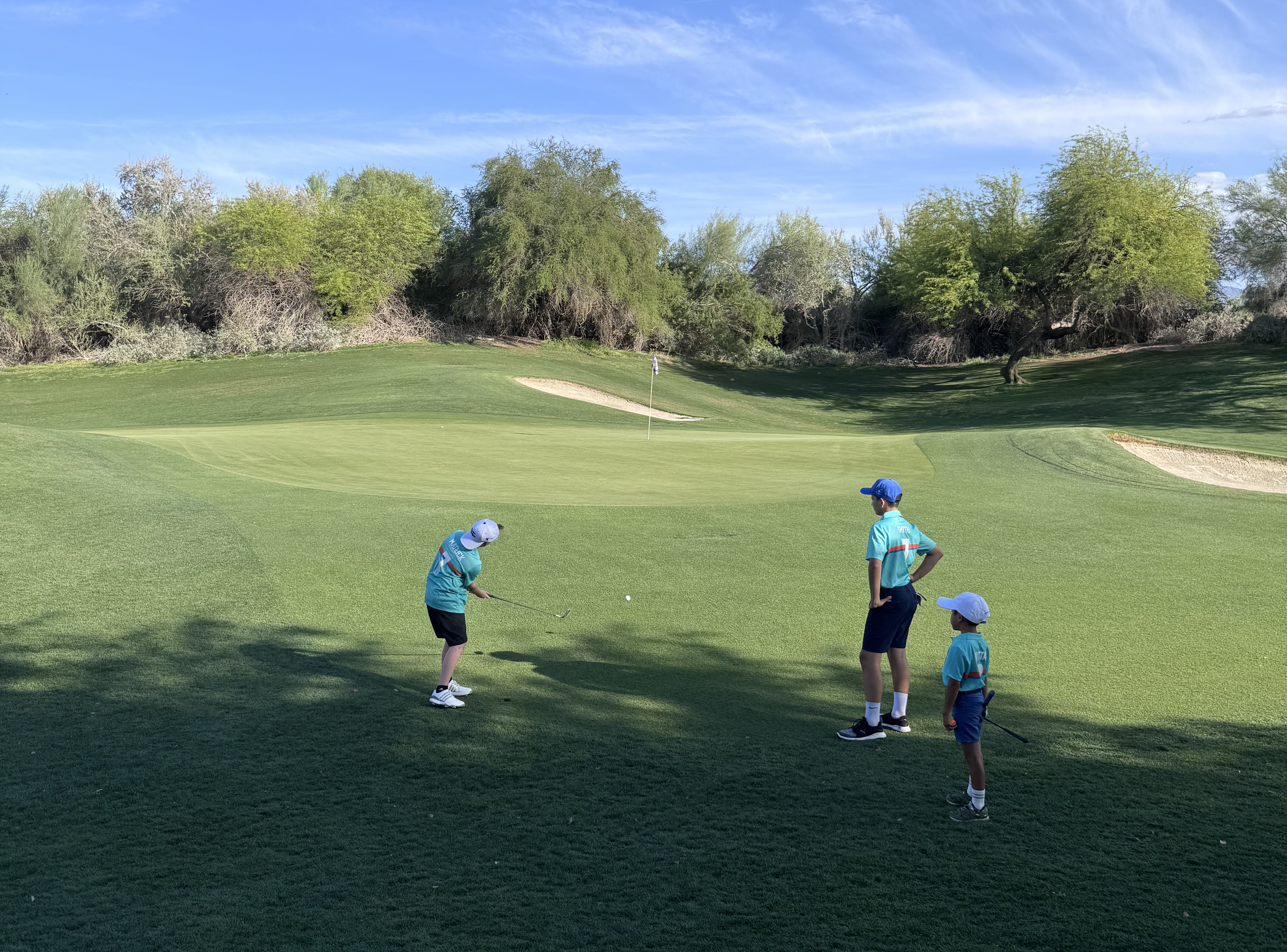 Three junior golfers practicing near a green at Desert Willow Golf Resort, with one mid-swing and two others observing. The course is surrounded by lush desert trees under a bright blue sky.